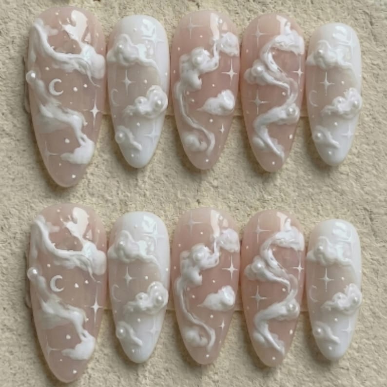 May include: Set of ten almond-shaped artificial nails. Each nail features a light pink base with white cloud and star designs. Some nails have a small letter "C" on them. The nails are arranged in two rows of five.