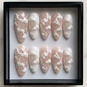 May include: A set of ten almond-shaped press-on nails. Each nail features a light pink base with white cloud and star designs. Some nails include a crescent moon. The text "CELESTIAL PRESS-ONS" is at the bottom.