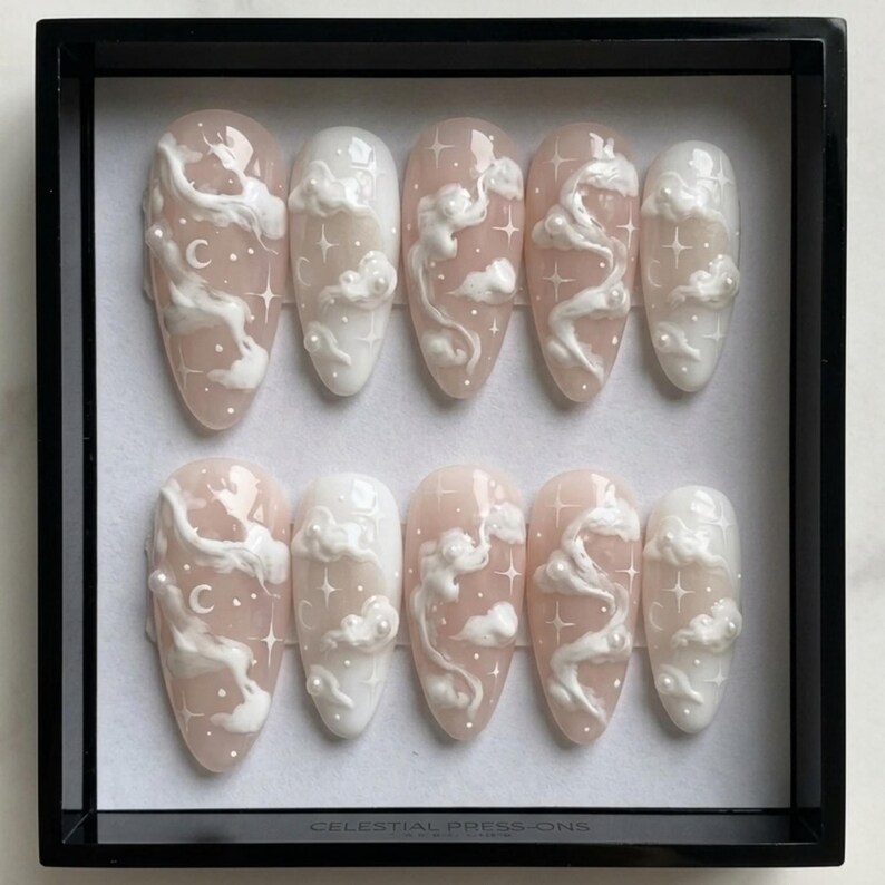 May include: A set of ten almond-shaped press-on nails. Each nail features a light pink base with white cloud and star designs. Some nails include a crescent moon. The text "CELESTIAL PRESS-ONS" is at the bottom.