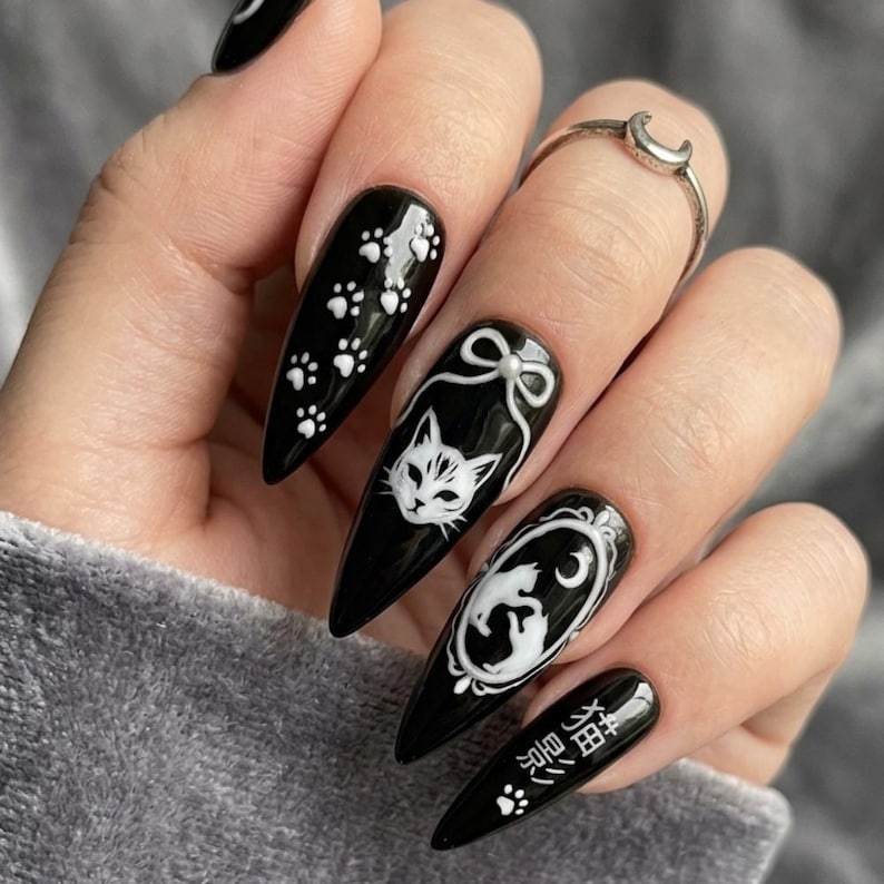 May include: Black stiletto nails with white cat-themed designs. One nail features paw prints, while others showcase cat faces, a cat silhouette in an oval frame, and Japanese text. A silver ring with a crescent moon is also visible.