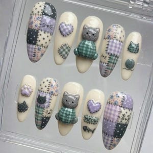 Könnte beinhalten: Ein Satz von zehn mandelförmigen Kunstnägeln mit Katzenmotiv. Designs umfassen Patchworkmuster in Lavendel, Grün und Creme, Katzenköpfe, Pfotenabdrücke, Herzen und 3D-Katzenfiguren. Einige Nägel zeigen das Wort "MEOW" und eine Fischgräte.
