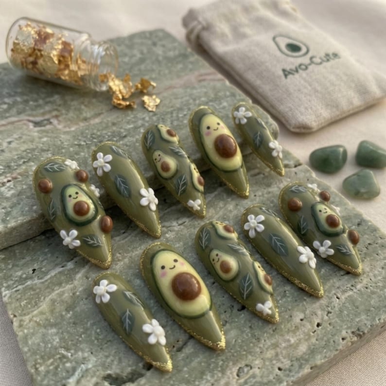 Pode incluir: Um conjunto de dez unhas artificiais longas em forma de am&ecirc;ndoa com um tema de abacate. Cada unha &eacute; pintada de verde-oliva com desenhos de abacate, flores brancas e detalhes de glitter dourado. Uma pequena bolsa com o texto "Avo-Cute" tamb&eacute;m &eacute; vis&iacute;vel.