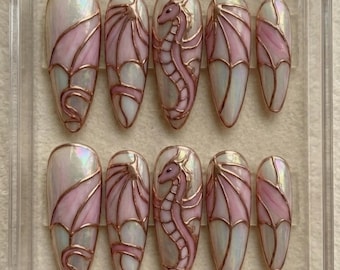 May include: A set of ten artificial nails with a pearlescent finish, featuring a pink dragon design with copper-colored outlines. The nails are arranged in a clear display case, showcasing the intricate artwork and design.
