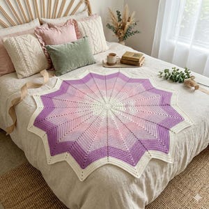 Könnte beinhalten: Eine sternförmige Häkeldecke mit einem Farbverlaufsmuster, das von einer cremefarbenen Mitte zu Nuancen von Hellrosa, Mauve und Dunkelviolett übergeht, abgeschlossen mit einem cremefarbenen Rand entlang der spitzen Kanten des Designs.
