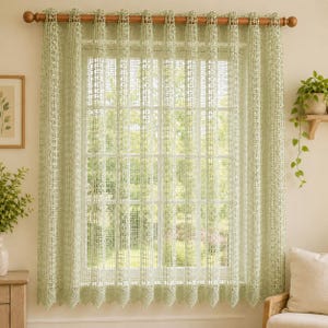 May include: A light sage green curtain featuring an intricate, open-weave crochet pattern with a grid-like texture. The curtain hangs from a wooden rod and displays a scalloped bottom edge, allowing soft light to filter through the fabric.