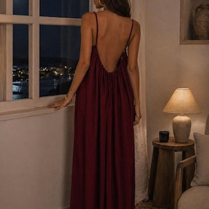 May include: A long, burgundy dress with thin straps and a low back. The dress has a flowing design, suitable for evening wear. The image is taken indoors, with a window and a view of a night scene.