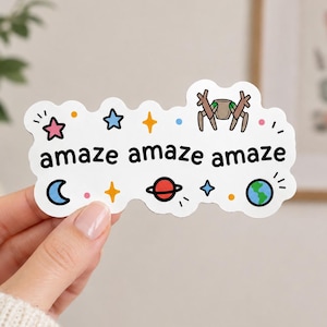 May include: A white die-cut sticker featuring the text "amaze amaze amaze" in black lowercase letters. The design includes colorful illustrations of a pink star, blue star, yellow sparkles, a crescent moon, a planet with rings, and the Earth.