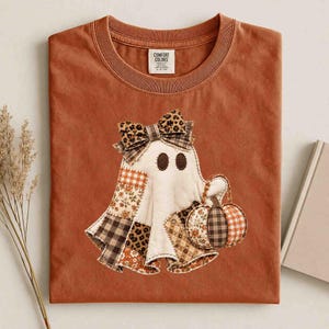 May include: A rust-colored t-shirt featuring a graphic of a ghost wearing a leopard print bow and holding a pumpkin. The ghost is decorated with various patchwork patterns including plaid, floral, and leopard print. The shirt has a Comfort Colors label.