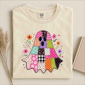 May include: A cream-colored t-shirt featuring a central graphic of a ghost made of various colorful patchwork squares, including floral, polka dot, plaid, and heart patterns, surrounded by small illustrated flowers and stars, with a Comfort Colors label.