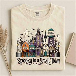 Puede incluir: Una camiseta color crema con un gráfico de un pueblo espeluznante que incluye una torre de agua con la etiqueta Petersburg, un huerto de calabazas y una tienda de pociones. El diseño incluye fantasmas, murciélagos y árboles desnudos, con el texto Spooky in a Small Town.