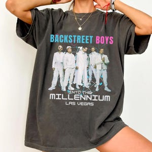 Op de afbeelding: Donkergrijs t-shirt met de tekst "BACKSTREET BOYS" in blauw en roze, en een groepsfoto van de band in witte outfits. Daaronder staat "INTO THE MILLENNIUM LAS VEGAS".
