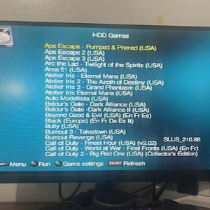 May include: A computer monitor displaying a blue menu screen titled HDD Games, featuring a list of video game titles including Ape Escape, Atelier Iris, and Call of Duty. The interface includes navigation options for Menu, Run, Game settings, and Refresh.