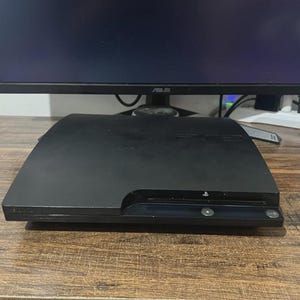 Peut inclure: Une console PlayStation 3 noire, une console de jeux vidéo, est posée sur une surface en bois. La console est rectangulaire avec une fente pour les disques et un bouton d'alimentation. Un écran d'ordinateur est en arrière-plan.