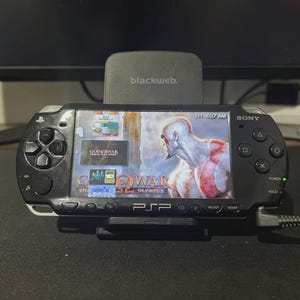 Może przedstawiać: Czarna konsola do gier przenośna Sony PSP z grą "God of War". Konsola jest zadokowana w czarnej stacji ładującej. Ekran pokazuje postać z czerwonymi oznaczeniami. Konsola posiada przyciski i joystick.