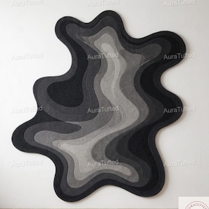 Könnte beinhalten: Ein großer, abstrakter Teppich mit einem geschichteten Design in Grau- und Schwarztönen. Der Teppich hat eine organische, fließende Form, mit dem Text "Aura Tufted" wiederholt an den Rändern. Das Design erzeugt ein Gefühl von Tiefe und Textur.