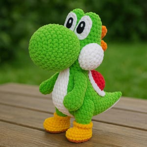 May include: A handmade crochet Yoshi doll, featuring a green body, white belly, and red and white shell. The doll has yellow boots and large, expressive eyes. The plush toy is displayed on a wooden surface.