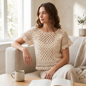 Könnte beinhalten: Ein cremefarbenes, kurzärmeliges Häkeltop mit rundem Ausschnitt. Das Oberteil hat ein detailliertes Lochmuster mit floralen Motiven und einen gewellten Saum. Die Frau sitzt auf einem hellfarbenen Sofa.