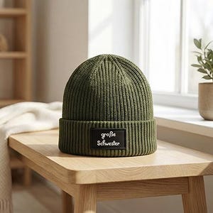 Könnte beinhalten: Eine olivgrüne Rippstrickmütze mit umgeschlagenem Saum. Ein schwarzes rechteckiges Patch auf der Vorderseite zeigt den Text "große Schwester" in Weiß. Die Mütze liegt auf einer hellen Holzoberfläche.