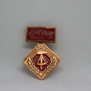 May include: A vintage medal with a rectangular top and a diamond-shaped bottom. The top is a textured, dark red, while the bottom features a gold-coloured design with a red centre and a hammer and compass symbol. The medal is likely a commemorative item.