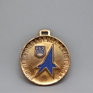 May include: A gold-colored circular medal with blue accents. The medal features the text "FÜR HERVORRAGENDE LEISTUNGEN" in a circular pattern around the edge. A blue star-like shape and the letters "FDJ" are in the center.