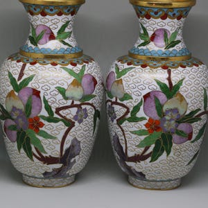 May include: A pair of matching cloisonné vases featuring a white background with a repeating cloud pattern. The vases are decorated with colorful depictions of peaches, green leaves, and small flowers, finished with gold-toned metal rims.