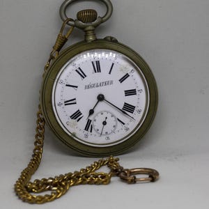 May include: A round pocket watch with a metallic brass-toned case and a white dial featuring black Roman numerals. The word REGULATEUR is printed on the face. It includes a sub-dial for seconds and a matching brass-toned metal chain with a circular link.