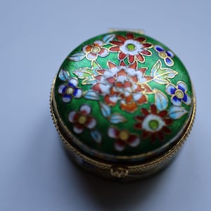 Pode incluir: Uma pequena caixa redonda para bugigangas com uma tampa de esmalte verde. A tampa é decorada com um padrão floral em vermelho, branco e azul, com detalhes dourados. A caixa tem uma base e uma borda de metal dourado.