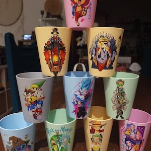 Puede incluir: Una colección de diez vasos de plástico cónicos apilados en forma de pirámide. Cada vaso presenta un estampado gráfico colorido y único que representa varios personajes animados, incluyendo figuras de anime e iconos de videojuegos.