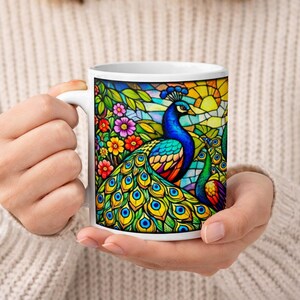 Może przedstawiać: Biały ceramiczny kubek z motywem pawia. Grafika przedstawia dwa pawie z niebieskimi, zielonymi i żółtymi piórami, na tle w stylu witrażu z kwiatami. Kubek jest trzymany w czyichś dłoniach.