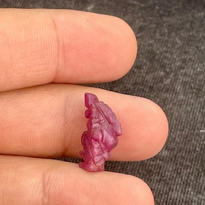 May include: A small, carved ruby gemstone in a deep red hue. The gemstone is held between two fingers and appears to be a miniature sculpture of a figure with detailed features. The carving has a textured surface.