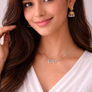 May include: A woman wearing a silver necklace with the Hindi word "राधा" and gold earrings. She has a red bindi on her forehead and is wearing a white top. Her dark hair is styled in waves.