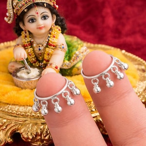 May include: Close-up of two fingers each adorned with a silver toe ring. Each ring features a series of small, dangling silver bells. In the background, a figurine of a deity is seated on a golden tray, wearing a jeweled crown and colorful garments.