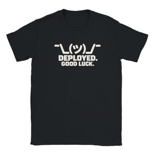May include: A black short-sleeved t-shirt featuring a white graphic print on the chest. The design includes a shrug emoticon above the text that reads, "DEPLOYED. GOOD LUCK." in a bold, sans-serif font centered on the front of the garment.
