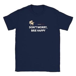 Peut inclure: Un t-shirt bleu marine à manches courtes avec un motif représentant un morceau de brie en dessin animé portant des lunettes de soleil. À côté du fromage se trouve une petite bande horizontale bleue, blanche et rouge. Texte : DON'T WORRY, BRIE HAPPY.