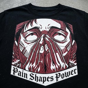 May include: Black crew neck t-shirt with a graphic illustration of a person's face, hands covering the face. The illustration is in shades of red and white. The text "Pain Shapes Power" is printed in a banner at the bottom of the graphic.