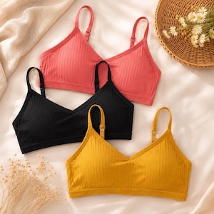 May include: A set of three ribbed bralettes in coral, black, and mustard yellow. Each bralette features a V-neckline, thin adjustable shoulder straps, and a wide elasticised underband. The fabric has a vertical ribbed texture throughout the design.