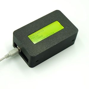 Puede incluir: Un dispositivo electrónico rectangular negro con una pantalla LCD verde que muestra texto blanco. El texto dice "Moduleenis P0010 LCD2004 I2C compatible". Un cable USB gris está conectado.
