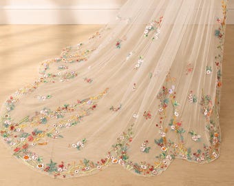 May include: A sheer, floor-length veil in a light cream color featuring intricate, multi-colored floral embroidery. The delicate fabric displays a variety of small flowers in shades of red, blue, yellow, and green, with a scalloped, embroidered edge.