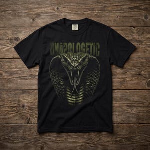 May include: A black short-sleeved t-shirt featuring a graphic of a cobra with its hood flared and mouth open, displaying fangs and a forked tongue. Above the snake, the word UNAPOLOGETIC is printed in a distressed, olive-green block font.