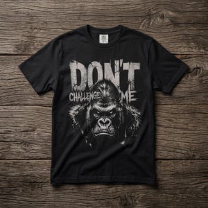 Puede incluir: Una camiseta negra de manga corta con un gráfico blanco de una cara de gorila centrada en el pecho. Sobre el gorila, el texto dice DON'T y debajo dice CHALLENGE ME en un estilo de fuente desgastado y envejecido de color blanco.