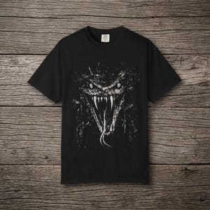 Könnte beinhalten: Ein schwarzes T-Shirt mit kurzen Ärmeln und einem kontrastreichen weißen Grafikdruck eines Schlangenkopfes mit gefletschten Zähnen und herausgestreckter Zunge. Das Design wirkt wie mit Tinte bespritzt. Ein kleines weißes Etikett befindet sich am Kragen.