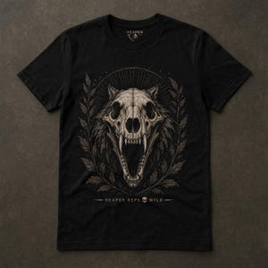 May include: A black short-sleeve t-shirt featuring a beige graphic of a wolf skull with an open jaw, surrounded by a circular laurel wreath and sunburst lines. The text REAPER REPS WILD with a small skull icon is printed below the graphic.
