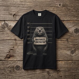 Könnte beinhalten: Ein schwarzes Kurzarm-T-Shirt mit einer Grafik eines Bibers vor einer Messlatte. Der Biber hält ein Schild mit der Aufschrift "UNAUTHORIZED CONSTRUCTION" in schwarzen Großbuchstaben. Die Messlatte zeigt Maße im imperialen System.
