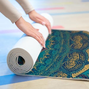 Op de afbeelding: Een witte yogamat wordt uitgerold en onthult een patroon in teal en goud. De mat ligt op een lichtgekleurd oppervlak. Het ontwerp bevat bloemen- en paisley-elementen, waardoor een decoratief en functioneel item voor yoga en beweging ontstaat.