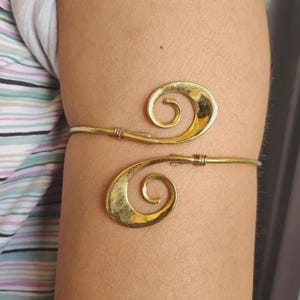 May include: A gold-coloured arm cuff featuring a spiral design. The cuff wraps around the arm with two decorative, swirling elements. The metal has a slightly textured appearance.