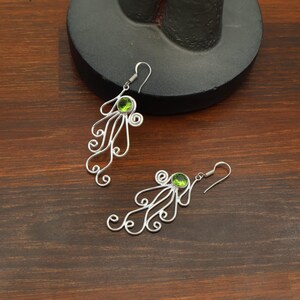 May include: A pair of silver-toned earrings with a unique design. Each earring features a green gemstone at the top, with swirling wire details resembling tentacles. The earrings are displayed on a wooden surface.