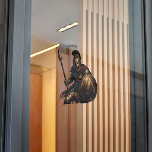 Könnte beinhalten: Ein Aufkleber mit einem spartanischen Krieger in Bronze-Rüstung, einem behelmten Kopf mit Kamm und einem roten Umhang, der einen langen Speer und einen runden Schild mit einem zentralen Lambda-Symbol hält. Der Krieger hat einen grimmigen Ausdruck.