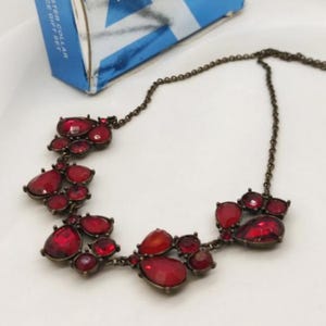 Op de afbeelding: Een ketting met een donkere metalen ketting en clusters van rode, druppelvormige edelstenen. De edelstenen variëren in grootte en zijn gezet in een donkere metalen zetting. Een blauw-witte doos staat op de achtergrond.