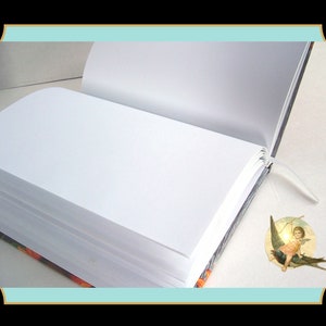 May include: An open hardcover book with blank white pages and a thin white ribbon bookmark. The book features a dark cover with orange floral patterns visible at the edges. A small circular sticker depicting a winged cherub is placed on the page.