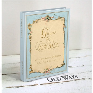 May include: A light blue wedding guest book with a gold and floral design. The cover reads "Guests & Well Wishes" and "Mr. and Mrs. Nathan McMurchy" with the date "June 18, 2019". The book is titled "Old Ways" on the bottom.